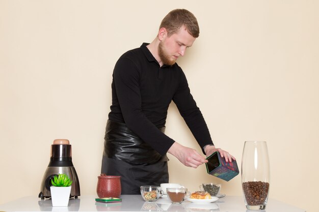 Joven barista en traje de trabajo negro con ingredientes y equipo de café semillas de café marrón sobre blanco