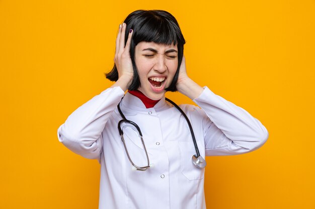 Joven bastante caucásica disgustada en uniforme médico con estetoscopio tapándose los oídos con las manos