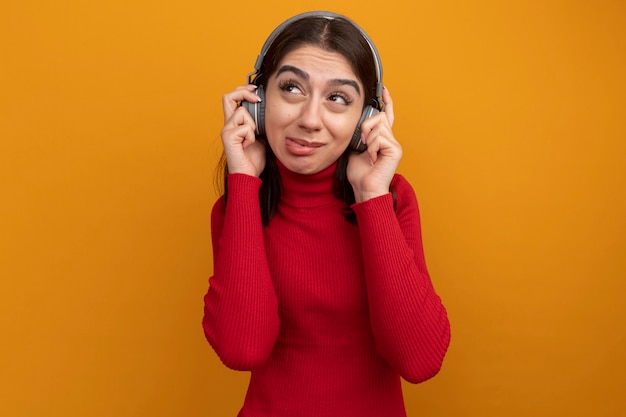 Joven bastante caucásica molesta vistiendo y agarrando auriculares mirando al lado mostrando la lengua