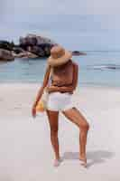 Foto gratuita joven bastante caucásica mujer en forma bronceada en ropa de punto en la playa tropical con sombrero de paja con bolsa de cuerda reutilizable ecología llena de frutas manzana uva naranja