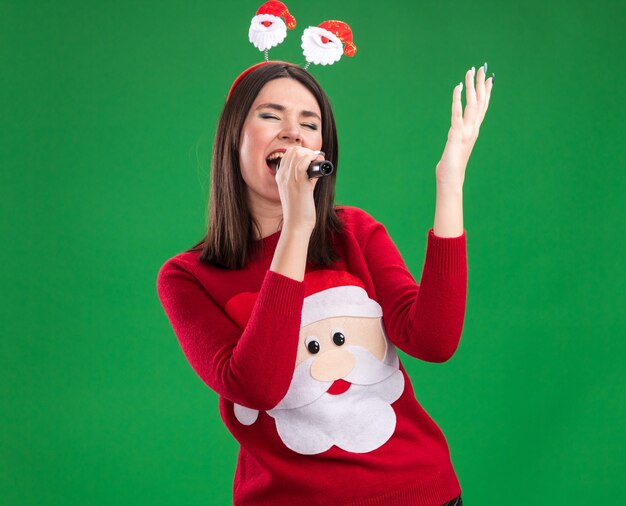 Joven bastante caucásica vistiendo suéter de santa claus y diadema sosteniendo micrófono levantando la mano cantando con los ojos cerrados aislado en la pared verde con espacio de copia