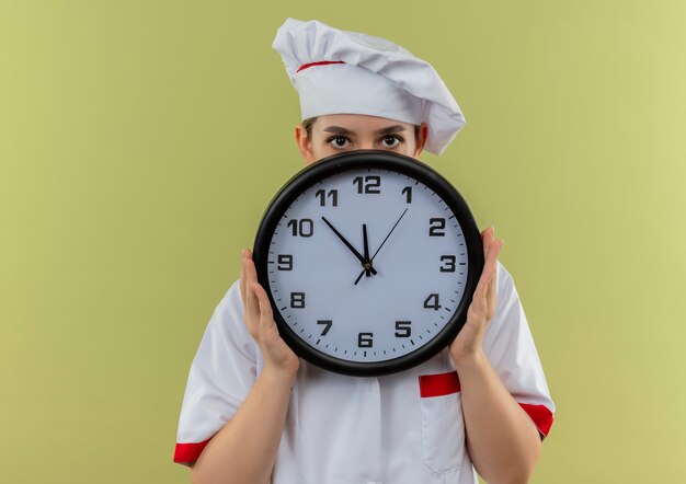 Joven bastante cocinero en uniforme de chef sosteniendo y escondido detrás del reloj aislado sobre fondo verde