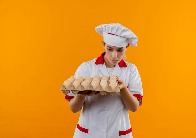 Joven bastante cocinero en uniforme de chef sosteniendo y mirando cartón de huevos