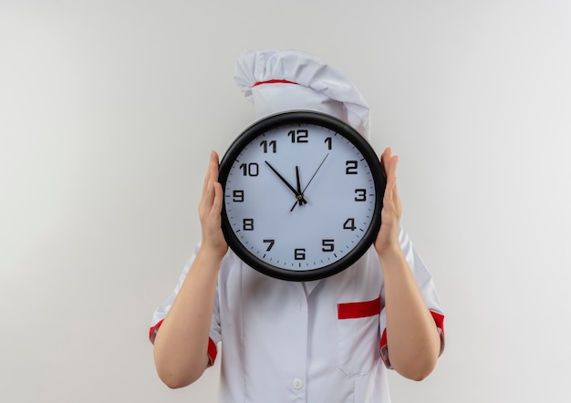 Joven bastante cocinero en uniforme de chef sosteniendo el reloj y escondiéndose detrás de él