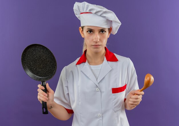 Joven bastante cocinero en uniforme de chef sosteniendo una sartén y una cuchara mirando