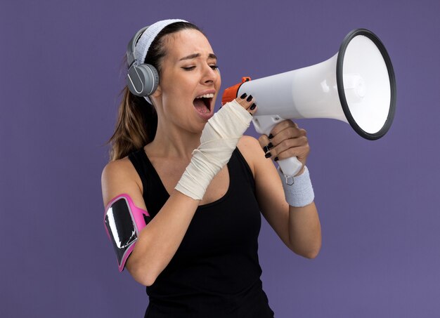 Joven bastante deportiva con auriculares y muñequeras de diadema y brazalete de teléfono con la muñeca lesionada envuelta con una venda hablando por el altavoz aislado en la pared púrpura