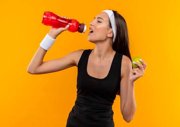 Joven bastante deportiva vistiendo diadema y pulsera bebiendo agua de la botella y sosteniendo apple