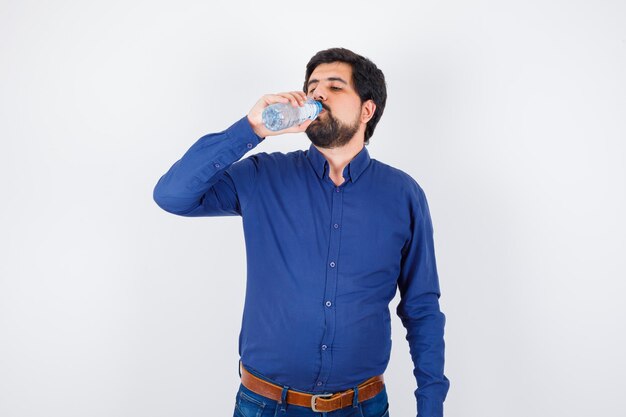 Joven bebiendo agua en camisa azul y jeans y mirando serio, vista frontal.