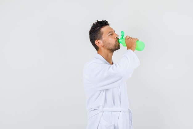 Joven bebiendo agua después del baño en bata de baño blanca, vista frontal.