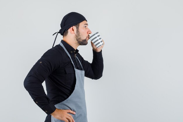 Joven bebiendo té o café en camisa, delantal y mirando enfocado. .