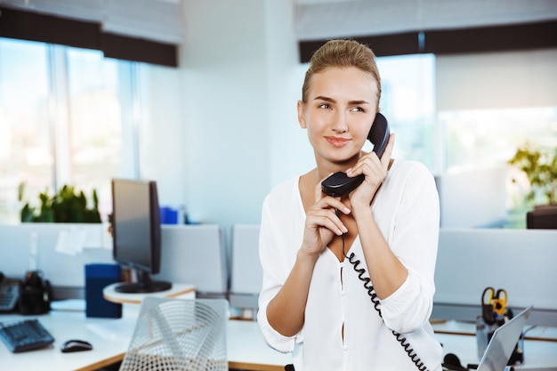 Joven y bella empresaria exitosa sonriendo, hablando por teléfono, sobre la oficina