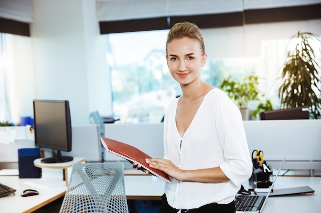 Foto gratuita joven y bella empresaria exitosa sonriendo, posando, sosteniendo la carpeta, sobre la oficina