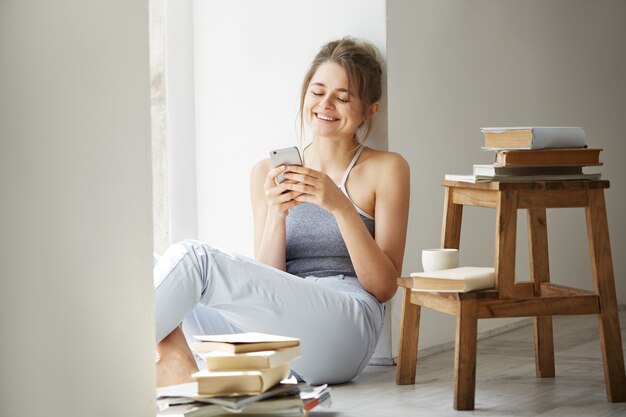 Joven y bella mujer adolescente navegando por la web mirando la pantalla del teléfono sonriendo sentado en el piso entre libros viejos cerca de la ventana sobre la pared blanca.