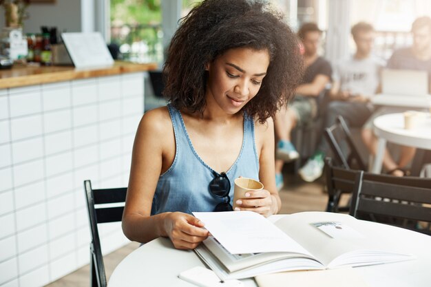 Joven y bella mujer africana estudiante sentado en la cafetería sonriendo mirando revista bebiendo café. Aprendizaje y educación.