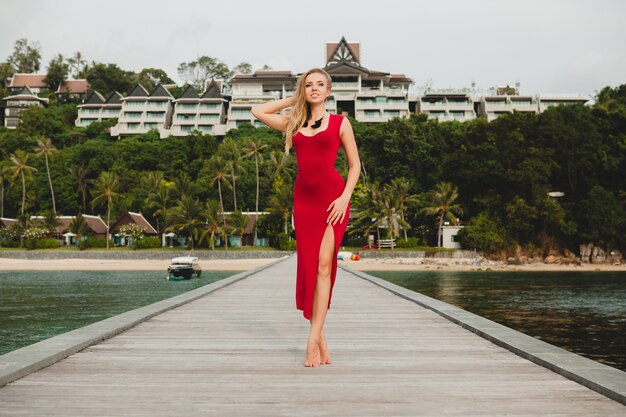 Joven y bella mujer atractiva sola en el muelle en el hotel resort de lujo, vacaciones de verano, vestido largo rojo, cabello rubio, ropa sexy, playa tropical, seductora, sensual, sonriente