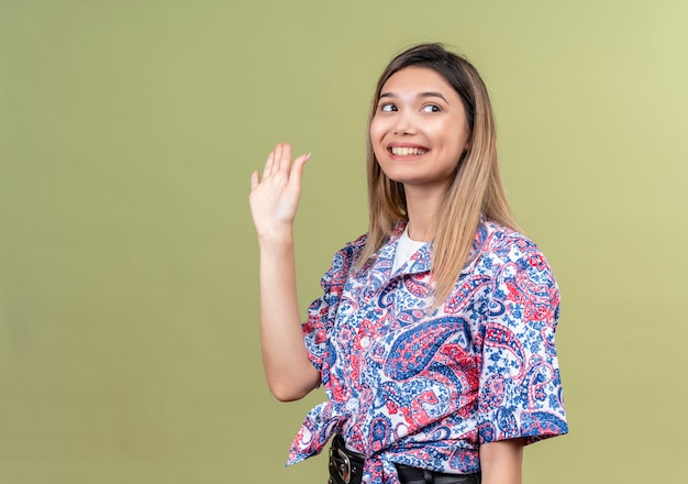 Una joven y bella mujer con camisa estampada de paisley agitando la mano y diciendo adiós mientras mira de lado en una pared verde