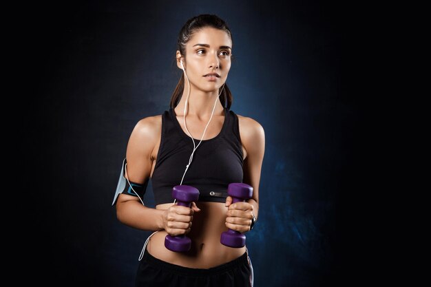 Joven y bella mujer deportiva bruntete entrenando con pesas sobre pared oscura