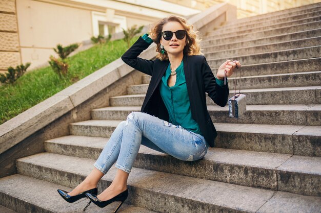 Joven y bella mujer elegante sentada en la calle, vestida con jeans, chaqueta negra, blusa verde, gafas de sol