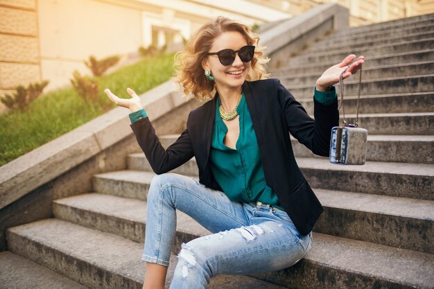 Joven y bella mujer elegante sentada en la escalera en la calle de la ciudad, vestida con jeans, chaqueta negra, blusa verde, gafas de sol, sosteniendo el bolso, estilo elegante, tendencia de moda de verano, sonriendo