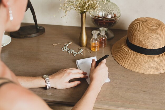 Joven y bella mujer elegante sentada en la mesa en la habitación del hotel resort, escribiendo una carta, sosteniendo la pluma, sombrero de paja, estilo vintage, primer plano de las manos, detalles, accesorios, diario de viaje