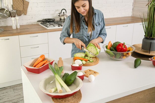 Una joven y bella mujer está preparando una ensalada de diversas verduras en la cocina.