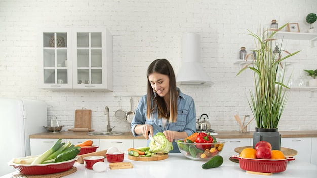 Una joven y bella mujer está preparando una ensalada de diversas verduras en la cocina.