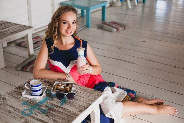 Joven y bella mujer con estilo en el café del mar, bebiendo batido de cóctel, gafas de sol, coqueta, estilo resort, traje de moda, sonriente, vestido de colores marinos