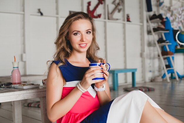 Joven y bella mujer con estilo en el café del mar, bebiendo capuchino caliente, estilo resort, traje de moda, sonriente, vestido de colores marinos, sentado en el piso, vacaciones, relajarse