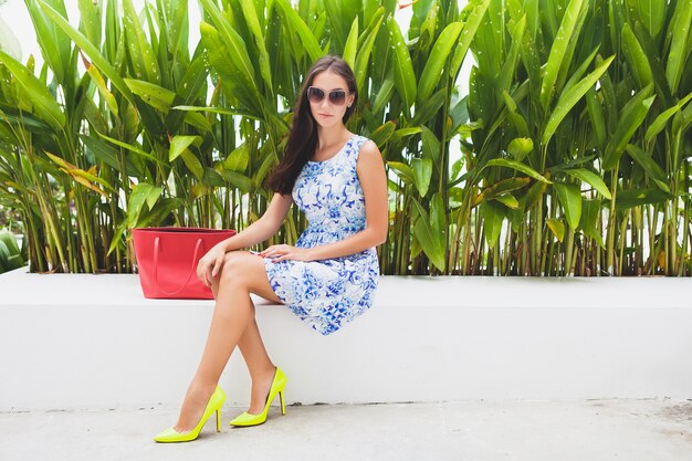 Joven y bella mujer con estilo en vestido estampado azul, bolso rojo, gafas de sol, estado de ánimo feliz, traje de moda, ropa de moda, sonriendo, sentado, verano, zapatos de tacón amarillo, accesorios