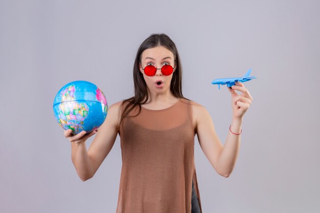 Joven y bella mujer con gafas de sol rojas con globo y avión de juguete sorprendido y sorprendido sobre la pared blanca