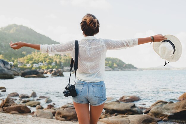 Joven y bella mujer hipster en vacaciones de verano en asia, relajándose en la playa tropical, cámara de fotos digital, estilo boho casual, paisaje de mar, cuerpo delgado y bronceado, viajar solo, libertad