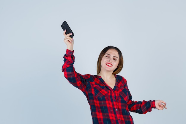 Joven y bella mujer levantando el teléfono, apuntando a la derecha en camisa casual y mirando feliz. vista frontal.