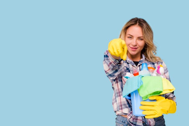 Joven y bella mujer limpiadora con cubo con productos apuntando a la cámara contra el fondo azul