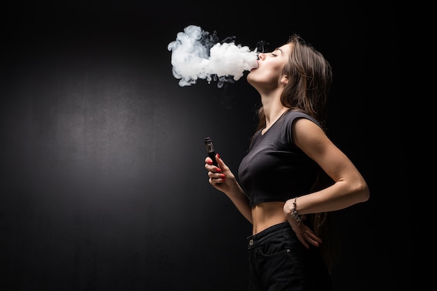Foto gratuita joven y bella mujer morena fumando, vaping cigarrillo electrónico con humo en la pared negra