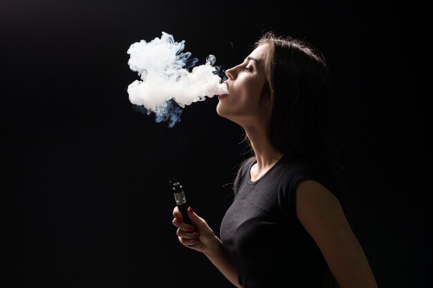 Joven y bella mujer morena fumando, vaping cigarrillo electrónico con humo en la pared negra