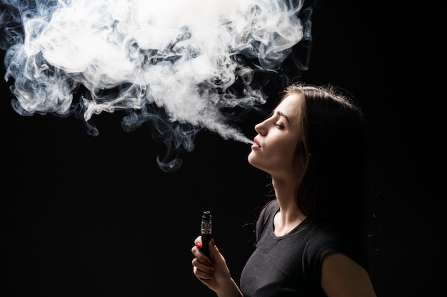 Joven y bella mujer morena fumando, vaping cigarrillo electrónico con humo en la pared negra