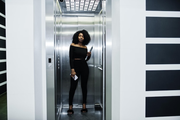 Foto gratuita joven y bella mujer de negocios afroamericana de moda con peinado afro en elegante soporte negro en el ascensor con teléfono y computadora portátil en las manos