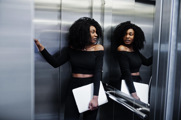 Joven y bella mujer de negocios afroamericana de moda con peinado afro en un elegante stand negro en el ascensor con una laptop en las manos