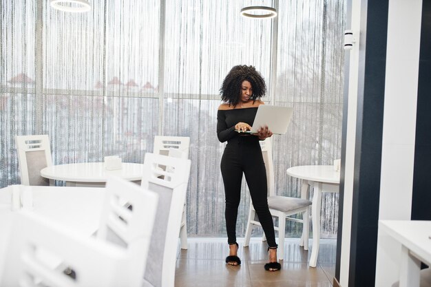 Joven y bella mujer de negocios afroamericana de moda con peinado afro en un elegante stand negro con una laptop en las manos