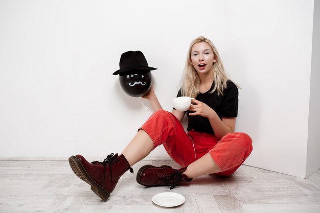 Joven y bella mujer rubia con globo negro con sombrero sentado en el piso tomando café sobre la pared blanca