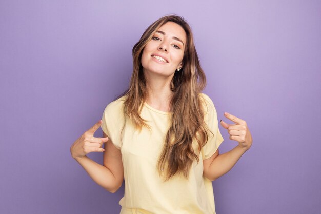 Joven y bella mujer satisfecha de sí misma en camiseta beige mirando confiada sonriendo apuntando a sí misma de pie sobre púrpura