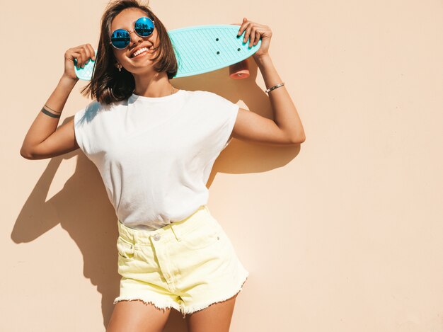 Joven y bella mujer sexy hipster sonriente en gafas de sol. Chica de moda en camiseta y pantalones cortos de verano. Mujer positiva con patín azul centavo posando en la calle cerca de la pared