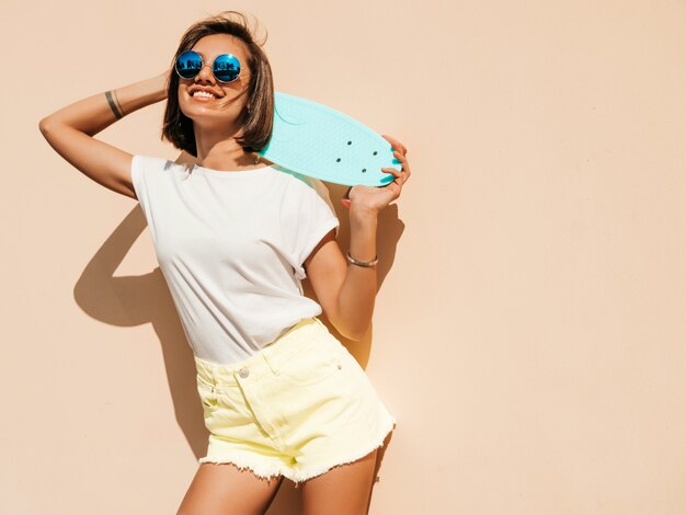 Joven y bella mujer sexy hipster sonriente en gafas de sol. Chica de moda en camiseta y pantalones cortos de verano. Mujer positiva con patín azul centavo posando en la calle cerca de la pared