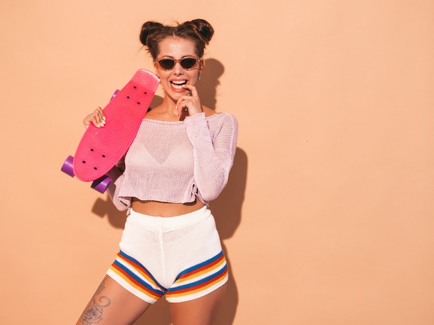 Joven y bella mujer sexy hipster sonriente en gafas de sol. chica de moda en verano tema punto cardigan, pantalones cortos. mujer positiva volviéndose loca con monopatín rosa centavo, aislado en la pared de color beige. dos hor