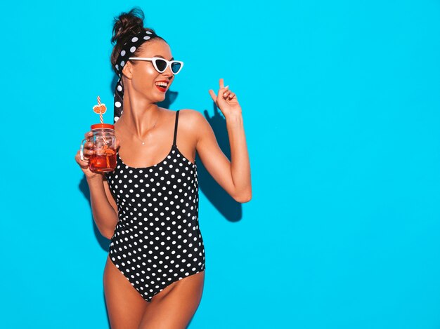 Joven y bella mujer sexy hipster sonriente en gafas de sol. Chica en traje de baño de verano guisantes traje de baño. Colocando cerca de la pared azul, bebiendo cóctel fresco bebida lisa. Muestra el signo de la paz