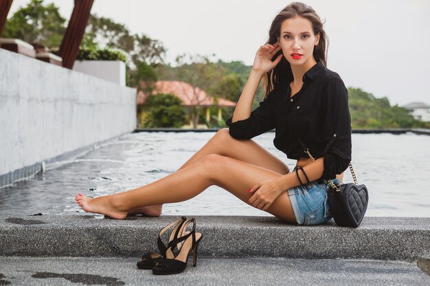 Joven y bella mujer sexy sentada en la piscina descalza, piernas bronceadas delgadas largas, zapatos de tacón alto, camisa negra, pantalones cortos de mezclilla, bolso, relajante, estilo de verano, vacaciones, detalles de primer plano