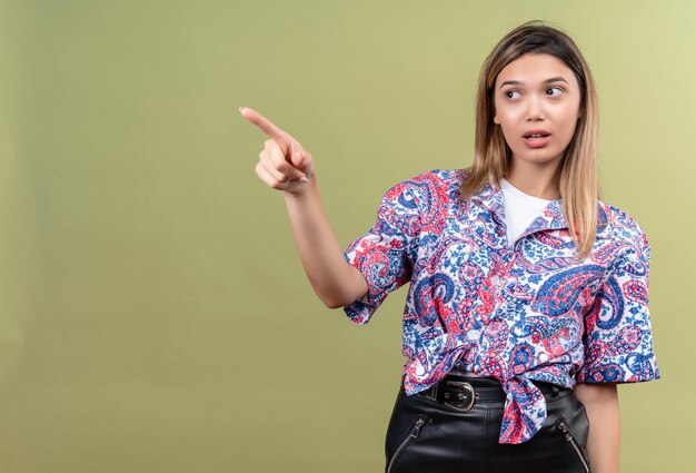 Una joven y bella mujer vistiendo camisa estampada de paisley apuntando con el dedo índice y mirando de lado en una pared verde