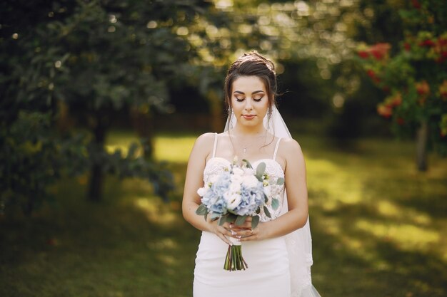 Una joven y bella novia está de pie en un parque de verano con ramo de flores