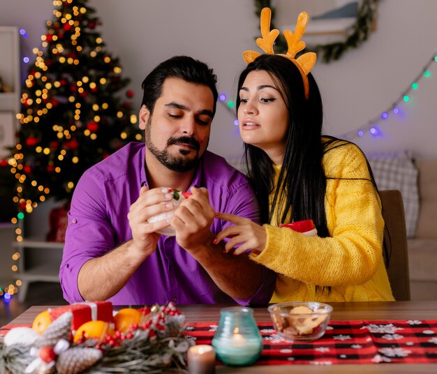 Joven y bella pareja hombre y mujer sentados a la mesa con tazas de té feliz en el amor en la habitación decorada de Navidad con árbol de Navidad en la pared