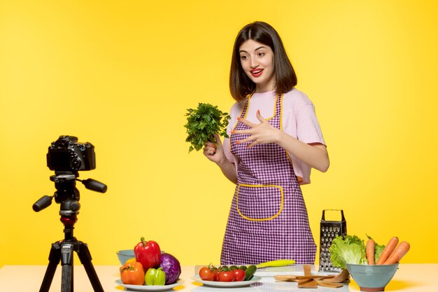 Joven bloguera de comida con delantal rosa grabando video para redes sociales sosteniendo un perejil